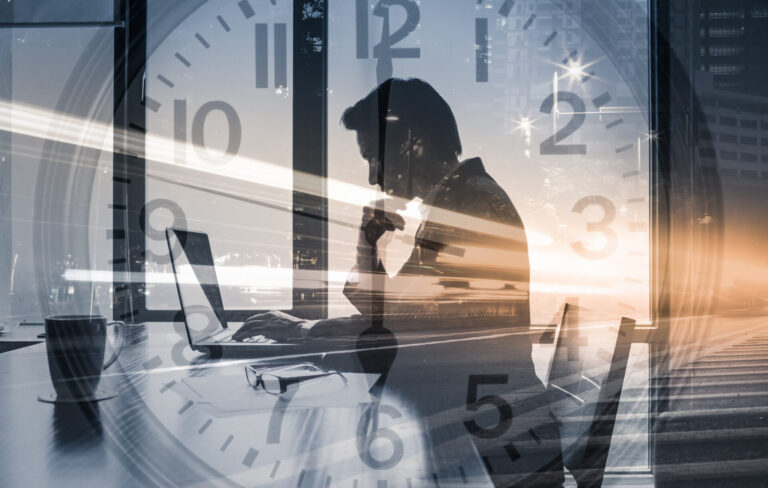 Ein Mitarbeiter sitzt an einem Schreibtisch. Überblendet wird eine tickende Uhr.